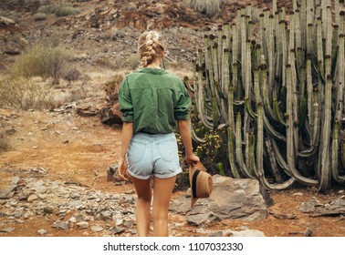 Young girl with long blonde hair wearing a shirt and jeans shorts is walking in the dry desert next to big cactus. Wild west style.