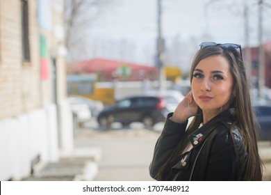 A Young Girl With Long Hair, European Descent, In A Leather Jacket, In The Spring, On A Sunny Day, Standing In The Parking Lot, Turned Over Her Shoulder In The Housing Estate, And Looks At The Camera