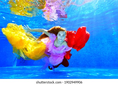 A Young Girl With Long Hair Swims And Poses Underwater In An Outdoor Pool With Colorful Fabrics In Her Hands. Fashion Portrait. Underwater Photography. Horizontal Orientation.