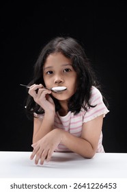 A young girl with long, wavy hair is sitting at a white table against a black background. The girl is holding a shiny metal spoon vertically in front of her face with both hands, and her lips are touc