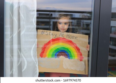 Young girl looking at infinity through a window and showing a picture of a rainbow on a cardboard, during coronavirus quarantine. Inspirational message “Stay at home”.