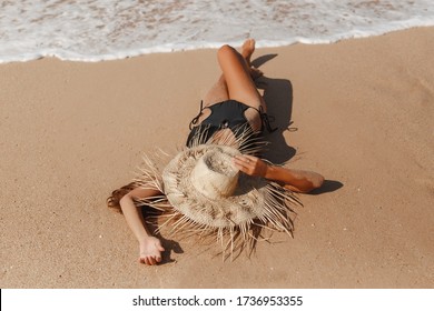 Young girl lying on white sand beach during her vacaion be the sea. She wears black bikini, cover the head with straw hat, waves touch her feets. Summer holidays concept