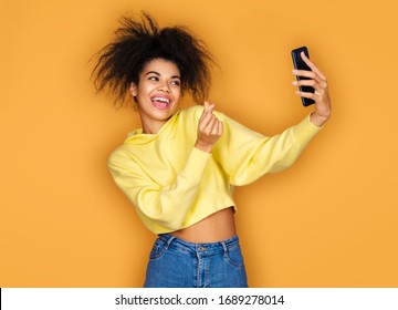 Young Girl Makes Korean Love Sign And Takes Selfie Photo, Looking At Phone. Photo Of African American Girl On Yellow Background