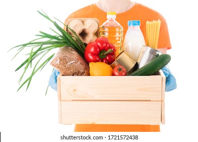 Young Girl In A Mask And An Orange T-shirt Holds A Wooden Box With Groceries For Delivery Isolated On White Background. Eggs, Can, Tomato, Oil, Milk, Pasta, Bottle, Lemon, Pepper, Cucumb