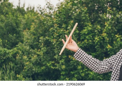 Young  Girl Musician Drummer In Plaid Shirt, Her Hand Twirling Drumstick With Virtuosity Against Dark Greenery Of Garden