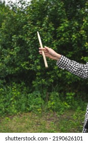 Young  Girl Musician Drummer In Plaid Shirt, Her Hand Twirling Drumstick With Virtuosity Against Dark Greenery Of Garden