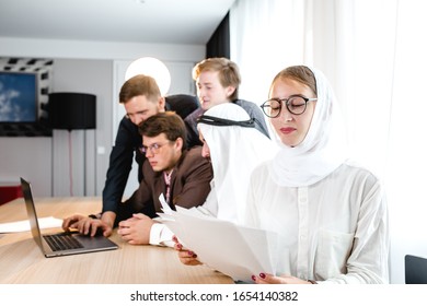  young girl in a national headscarf is holding sheets of paper at a meeting, in the background a meeting with an Arab representative and looking at a laptop.