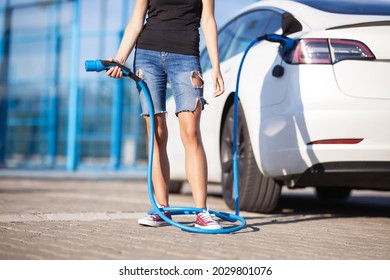 Young Girl Next To An Electric Car. Clumsy Wrapped With A Charging Cable Around Her Legs.