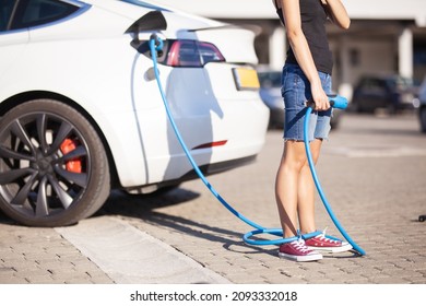 Young Girl Next To An Electric Car. Clumsy Wrapped With A Charging Cable Around Her Legs.