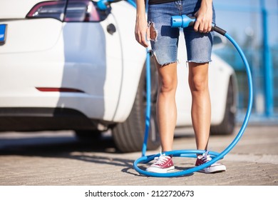 Young Girl Next To An Electric Car. Clumsy Wrapped With A Charging Cable Around Her Legs.