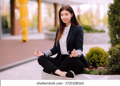 Young Girl, Office Worker, With Long Brunette Hair In Dark Blue Jacket And Striped Shirt Sits In A Pose Of Yoga Practice Outside Near Office Building And Smiles Looking At The Camera