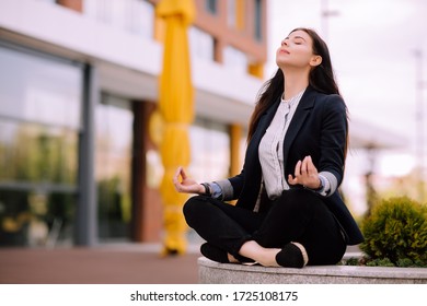 Young Girl, Office Worker, With Long Brunette Hair In Dark Blue Jacket And Striped Shirt Sits With Closed Eyes And  
Breathe Deeply In A Pose Of Yoga Practice Outside Near Office Building