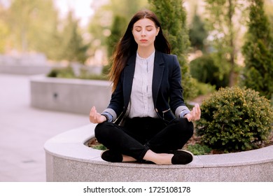 Young Girl, Office Worker, With Long Brunette Hair In Dark Blue Jacket And Striped Shirt Sits With Closed Eyes In A Pose Of Yoga Practice Outside Near Office Building 