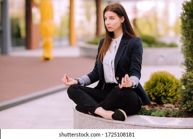 Young Girl, Office Worker, With Long Brunette Hair In Dark Blue Jacket And Striped Shirt Sits In A Pose Of Yoga Practice Outside Near Office Building And Looks In Front Of Her