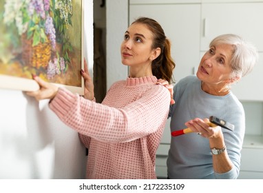 Young Girl And Older Woman Hanging Together Painting With Depicted Bouquet Of Lilacs On Wall Indoors .