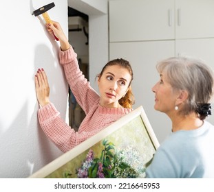 Young Girl And Older Woman Hanging Together Painting With Depicted Bouquet Of Lilacs On Wall Indoors .