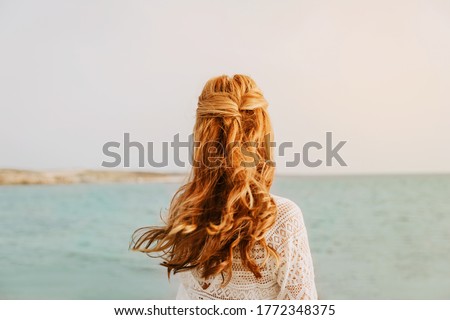 Young girl on the beach. With red long hair, standing with her back. Wedding hairstyle. Sea wedding concept. 