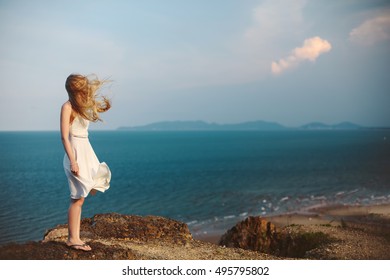 Young Girl On Rock Beach, Wind In Blond Hair And White Dress