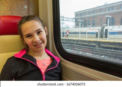 Young Girl On A Train Getting Ready For A Trip. Girl Smiling At Camera With Her Back To A Window On A Cloudy, Cold Day At A Train Station.