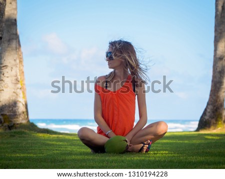 young girl on vacation on a tropical island, sitting on the green grass near the green coconut. ocean background.