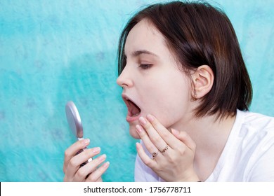 A Young Girl With An Open Mouth Looks At A Sore Tooth Through A Small Mirror. Turquoise Background, Close-up.