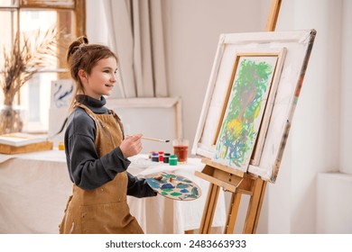 A young girl is painting a picture on a canvas. She is wearing an apron and has a brush in her hand. The painting is colorful and lively, with a mix of green and blue hues - Powered by Shutterstock