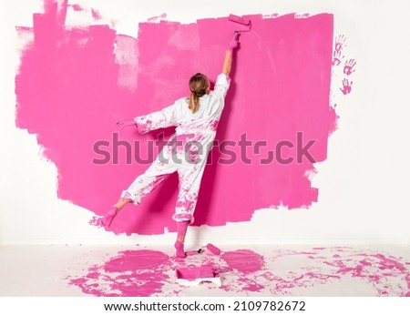 Young girl painting a wall with pink color. She is wearing working clothes and both her feet are dipped in pink color.