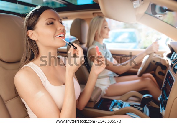 Young Girl Paints Lips in Car Interior. Friend\
Drives Car behind Wheel. Lifestyle Concept. Travel Concept. Summer\
Travel Concept. Summer Vacation Concept. Drive by Car. Pretty\
Caucasian Driver.
