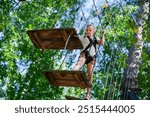 Young girl participating in outdoor adventure park, navigating through a rope obstacle course. She is wearing safety harness and carefully crossing wooden platform suspended between ropes