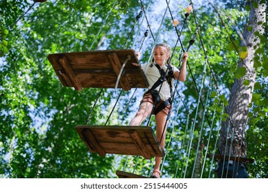 Young girl participating in outdoor adventure park, navigating through a rope obstacle course. She is wearing safety harness and carefully crossing wooden platform suspended between ropes - Powered by Shutterstock