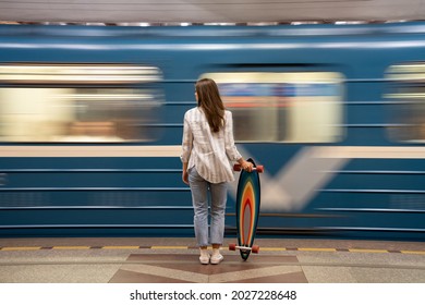 Young girl passenger with longboard standing on subway station platform with blurry moving blue train on background, rear view. Woman with skateboard watching metro pass fast at the station.