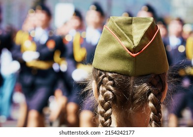 A young girl with pigtails at the military parade on Victory Day. Close view from behind. In the background, a column of female servicemen is marching in blur. Woman and war concept. Sunny day. - Shutterstock ID 2154242177