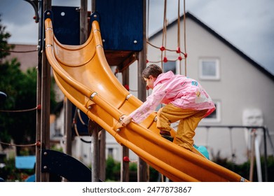 Young girl in a pink raincoat climbing up an orange slide at a playground on a rainy day. The scene captures her determination and playful spirit. - Powered by Shutterstock