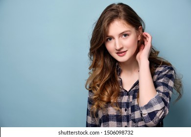 A Young Girl In A Plaid Shirt Is Standing Against A Blue Background And Looking At The Camera, Brushing Her Hair Behind Her Ear.