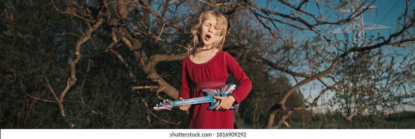 Young Girl Playing Guitar Toy Outdoor. Girl Pretending To Be Rock Music Star. Adorable Funny Child Having Fun In Park. Funny Authentic Candid Childhood Moments. Web Banner Header.