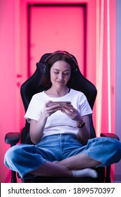 Young Girl Playing Mobile Online Game On A Smart Phone While Sitting On Gaming Chair In Colorful Neon Light Room.