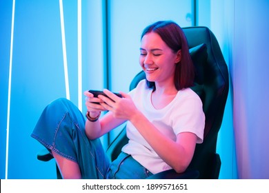 Young Girl Playing Mobile Online Game On A Smart Phone While Sitting On Gaming Chair In Colorful Neon Light Room.