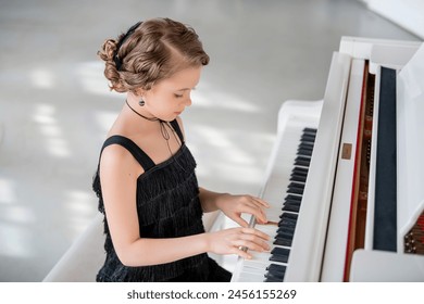 A young girl is playing the piano in a black dress. She is smiling and she is enjoying herself. The piano is white and has a black keyboard - Powered by Shutterstock