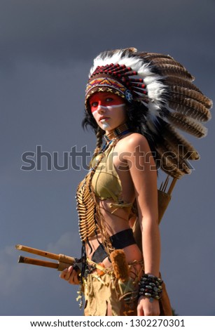 A young girl plays the part of a native American Indian 
girl. Dressed as a native Indian wearing a feathered 
headdress. She poses outdoors in a nature surrounding.