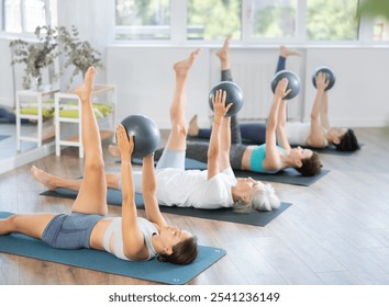 Young girl practicing Pilates with group of women in bright studio, lying on exercise mat with stability ball with extended arms and lifting one leg, focusing on core strength, balance, and - Powered by Shutterstock