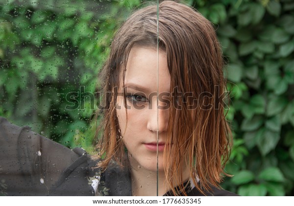 A young girl with predatory look and wet hair is\
looking at the camera. Wet glass with drops of water divides her\
face in half.