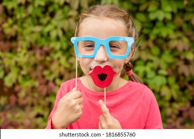 Young Girl Is Preparing For Birthday Party With Photo Booth Props. Photobooth Birthday And Party Set And Wedding.Happy Child With Glasses, Hat, Lips Props At Party Outdoor, On Leaves Background