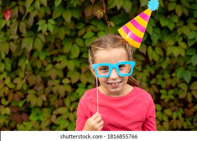 Young Girl Is Preparing For Birthday Party With Photo Booth Props. Photobooth Birthday And Party Set And Wedding.Happy Child With Glasses, Hat, Lips Props At Party Outdoor, On Leaves Background