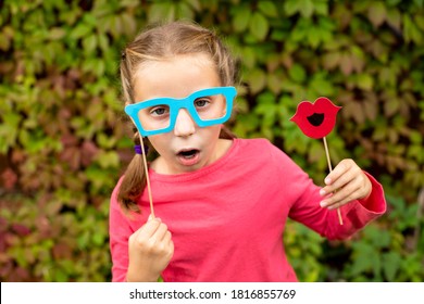 Young Girl Is Preparing For Birthday Party With Photo Booth Props. Photobooth Birthday And Party Set And Wedding.Happy Child With Glasses, Hat, Lips Props At Party Outdoor, On Leaves Background