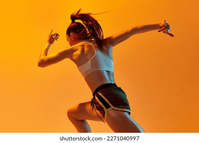 Young girl, professional runner athlete in uniform training, running over orange studio background in neon light. Concept of sportive lifestyle, health, endurance, action and motion. Ad