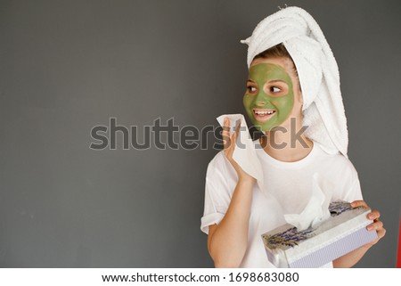 the young girl put a green mask on her face and with a towel on her head and takes care of herself. girl on a gray background smiling