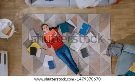 Young Girl in Red Coat and Blue Jeans is Lying Down on a Floor, Reading a Notebook. She Smiles and Laughs. Cozy Living Room with Modern Interior with Carpet, Workbooks and Backpack. Top View.