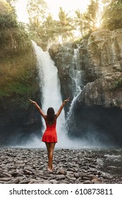 Young girl in red dress enjoying vacation and exploring beautiful waterfall at the sunset time during her luxury travel, wanderlust, globetrotter