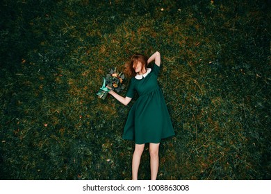 Young girl with red hairlying on ground. Portrait from above of cute teenager female relaxing on grass in summer. Lovely skinny female in beautiful stylish dress holding bouquet of flowers in hand. 