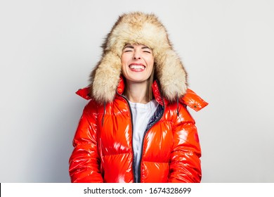 young girl in a red jacket and a fur hat, laughs with squinted eyes on a light background. Concept of laughter, good mood
